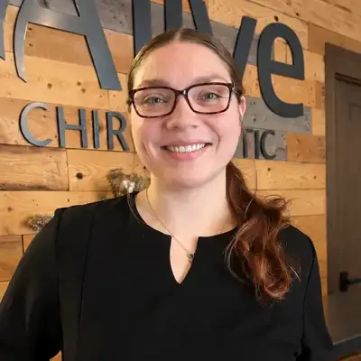 Team member wearing glasses and a black top standing in front of a wood-paneled wall with the Alive Chiropractic logo displayed in the background
