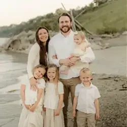 Family of five standing together on a sandy shoreline near the water, wearing light-colored clothing, with grassy dunes and driftwood in the background. Natural outdoor portrait taken by the beach.